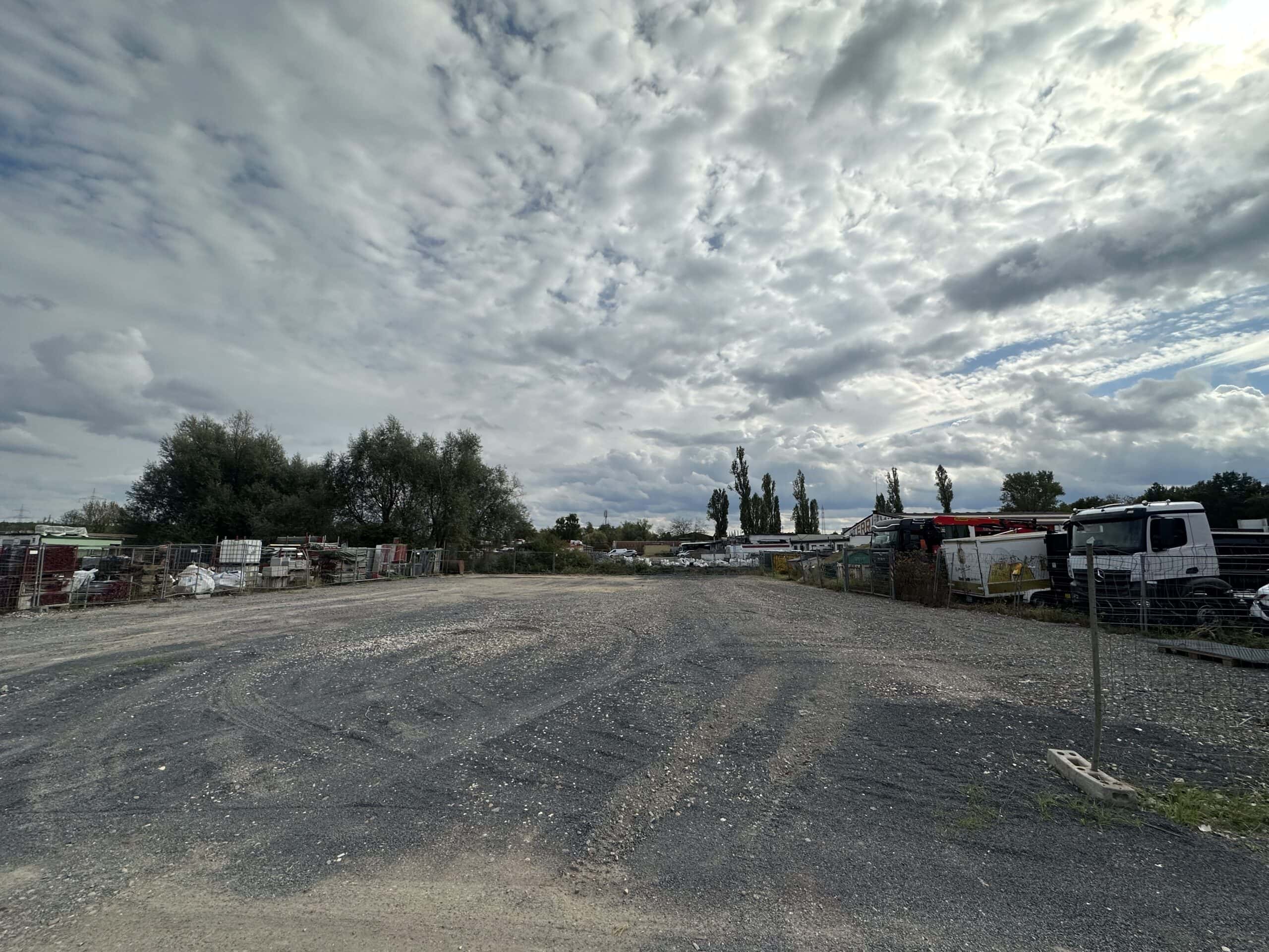 Weitläufiger Schrottplatz mit Kiesplatz, Wolken am Himmel, Bäume und Fahrzeuge im Hintergrund.